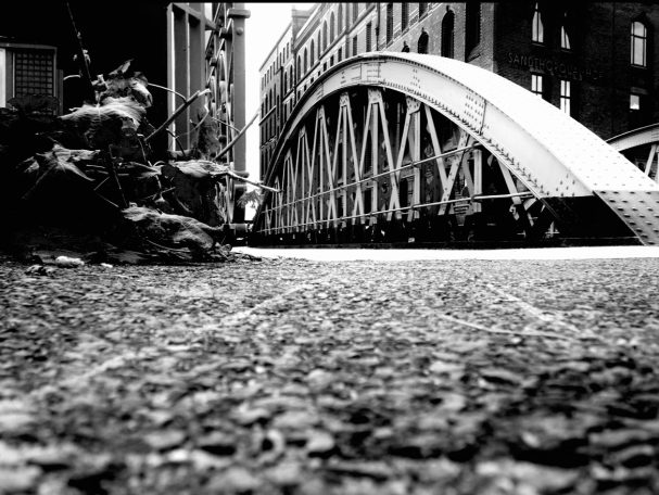 "Schwarzweißfoto aus Bodensicht: Blick auf eine Stahlbrücke zwischen alten Gebäuden