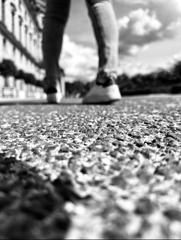 Schwarzweißaufnahme eines Fußgängers von unten, Fokus auf dem Strukturierten Asphalt im Vordergrund