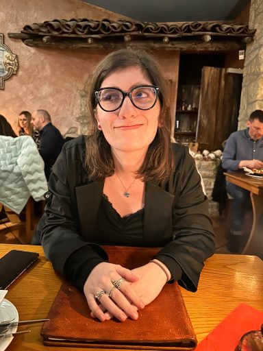 Frau mit Brille sitzt an einem Holztisch im ein Rustikalen Restaurant und schaut ruhig in die Kamera. Gäste im Hintergrund.