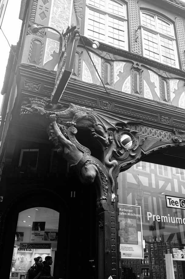 Schwarzweißfoto eines Fachwerkgebäudes mit großen Fenstern in Marburg, Ein Drachenornament sitzt oben links über dem Fensterrahmen.